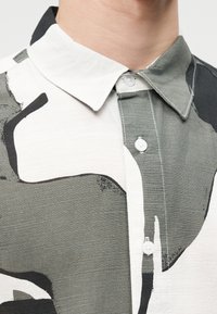 Prim-plan cu o persoană purtând o cămașă cu nasturi, având un model abstract în negru, alb și gri, și un guler ascuțit.