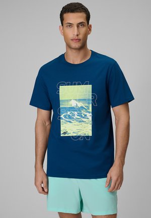 Mężczyzna noszący ciemnoniebieską koszulkę z żółtą falowaną grafiką i napisem „SUMMER SEASON”, w połączeniu z jasnoturkusowymi szortami.