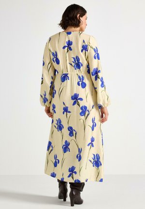 Donna vista di spalle che indossa un lungo vestito beige con stampa floreale blu e maniche lunghe, abbinato a stivali con tacco marrone scuro.