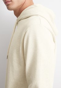 Felpa con cappuccio crema dal texture morbida, con coulisse e vestibilità comoda. La vista laterale evidenzia il design senza cuciture delle spalle.