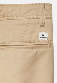 Beige Baumwollhosen mit einer Gesäßtasche, die ein kleines schwarz-weißes Markenlabel und sichtbare Nähte aufweist. Glatte Textur.