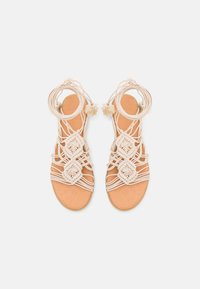 Sandali piatti beige con intricate cinghie in macramè, legacci per la caviglia e dettagli con frange, esposti su uno sfondo bianco.