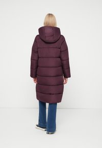 Dlhé dámske bundy s kapucňou v tmavo purpurovej farbe, so horizontálne prešitými panelmi a mierne obtiahnutým pásom, nosené s rozšírenými džínami.