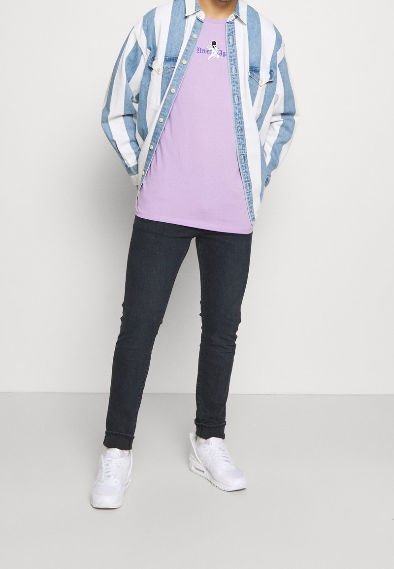Personne portant une veste en jean rayée bleu et blanc sur un t-shirt violet avec texte, un jean noir slim et des baskets blanches sur un fond uni.