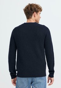 Temno modra pletena jopica z rebrastim vzorcem, okroglo ovratnikom in dolgimi rokavi, z oprijetim silhueto in subtilnimi šivi.