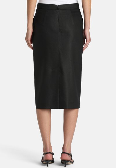 Kvinde iført en sort midi blyantsnørk med lynlås bagpå og sorte sandaletter med hæl, stående foran en ensfarvet hvid baggrund.