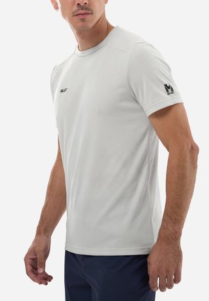 Homme portant une chemise à manches courtes gris clair ajustée de la marque Millet et un pantalon foncé, debout les bras détendus le long du corps, légèrement tourné vers la droite.