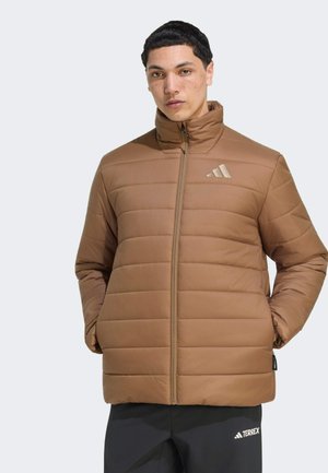 Uomo che indossa una giacca trapuntata marrone con collo alto e pantaloni neri, in piedi con le mani in tasca su uno sfondo chiaro e uniforme.