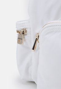 Bolsa de nylon blanca con cremalleras y herrajes en tono dorado, incluyendo un distintivo logo 'GUESS' en el tirador de la cremallera. Textura suave, diseño compacto.