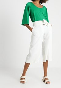 Groene top met korte mouwen gecombineerd met witte, brede culottes met een tailleband. Model draagt lichte sandalen. Schoon materiaal, casual ontwerp.