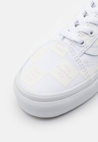 White canvas sneaker with a textured surface, featuring embossed square patches with words. Rubber sole provides grip and stability.