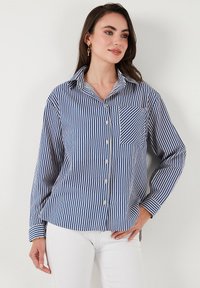 Camicia a righe blu e bianca in cotone. Maniche lunghe, colletto, taschino sul petto e orlo leggermente curvato. Texture liscia.