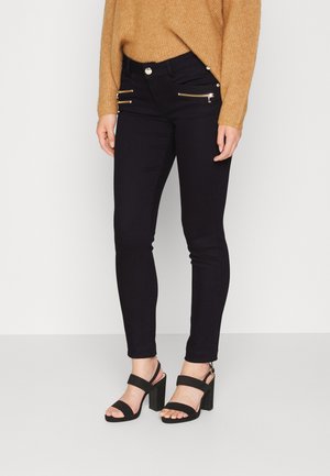 Jeans skinny negros con bolsillos con cremallera dorada, combinados con un suéter texturizado en color beige y sandalias negras de tacón. Tela suave, diseño ajustado.