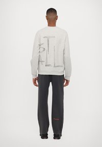 Grå sweatshirt med stor penseldragstext på ryggen, i kombination med mörkgrå byxor med röd brodyr nära fållen.