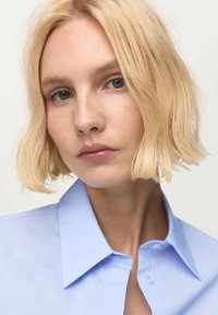 Svetlomodrá košeľa s golierom a hladkou textúrou. Osoba má krátke, vlnité blond vlasy a minimálne líčenie, čo zachováva prirodzený vzhľad.