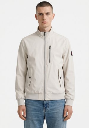 Młody mężczyzna stoi zwrócony przodem, ubrany w jasnobeżową kurtkę zapinaną na zamek z kieszenią na piersi na zamek oraz dwoma kieszeniami na patki zapinanymi na guziki.