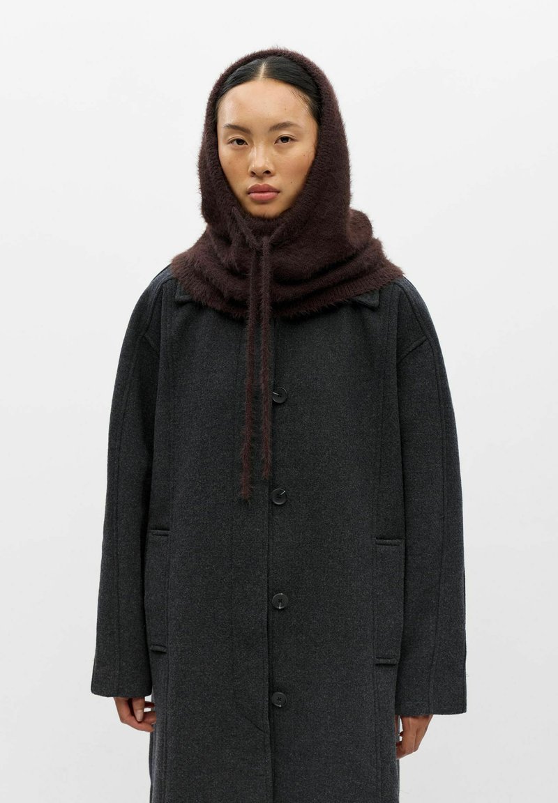 Jeune femme portant un manteau gris foncé boutonné et une épaisse capuche en tricot marron avec des cordons sur la tête.