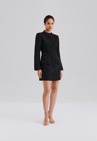 Schwarzes, strukturiertes Kleid mit langen Ärmeln, tailliertem Oberteil und zwei vorderen Taschen. Kombiniert mit silbernen Riemchensandalen.