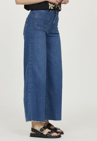 Jean en denim bleu à jambes larges avec un ourlet brut, comportant deux poches arrière et une taille haute, associés à des sandales noires.