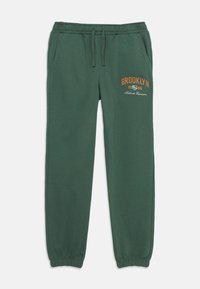 Grüne Baumwoll-Sweatpants mit elastischem Bund und Bündchen, mit braunem "BROOKLYN 1986" Grafik auf der Vorderseite und seitlichen Taschen.