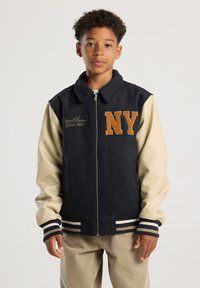 Boy wearing a black and beige varsity jacket with "NY" in orange letters, standing against a plain light background.