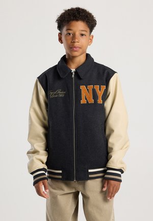 Ragazzo che indossa una giacca varsity nera e beige con la scritta "NY" in lettere arancioni, in piedi contro uno sfondo chiaro e semplice.