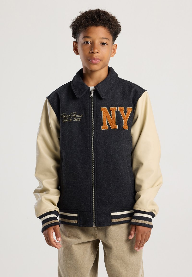 Boy wearing a black and beige varsity jacket with "NY" in orange letters, standing against a plain light background.