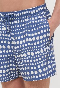 Svømmeshorts i blått stoff med et mønster av store hvite prikker, elastisk liv med snor og sidelommer.