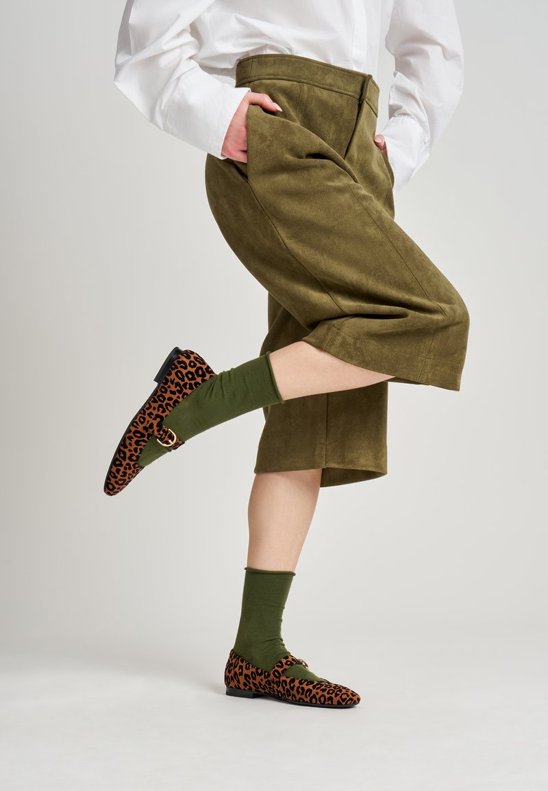 Mocasines de estampado de leopardo con patrón marrón y negro, combinados con calcetines verdes, llevados con pantalones de pernera ancha en verde oliva y camisa blanca.