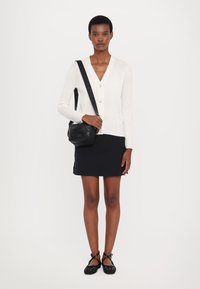 Bílý pletený cardigan s véčkovým výstřihem a zapínáním na knoflíky, zkombinovaný s černou sukní. Doplňky zahrnují černou crossbody tašku a polka dot baleríny.
