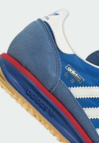 Modré semišové a textilní tenisky s bílými pruhy, bílou patou, červenými akcenty a texturovanou gumovou podrážkou. Obsahují zlatý branding.