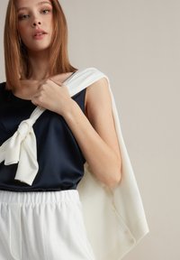 Granatowa satynowa bluzka bez rękawów połączona z białym dzianinowym swetrem narzuconym na ramiona, z detalem w postaci supełka i spodniami z elastycznym pasem.