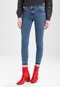 Kvinna som bär blå skinny jeans och blanka röda ankelstövlar med klack, i kombination med en svart tröja med snörade ärmar.