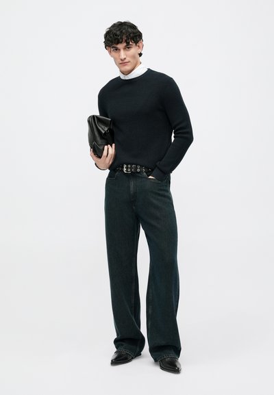 Suéter negro de canalé, jeans de pierna ancha de mezclilla oscura, cinturón de cuero negro con cortes circulares, zapatos de cuero negro y un bolso de mano negro.