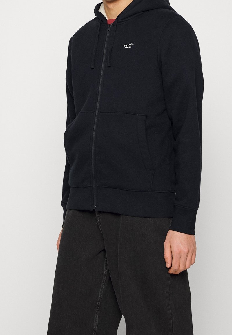 Svart zip-up hoodie tillverkad av mjukt tyg, med en frontdragkedja, två sidofickor och en liten logotyp på vänster bröst. Huvan ingår.