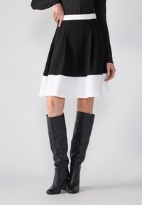 Jupe A-line en noir et blanc avec un design plissé, pr�ésentant une taille contrastante en blanc. Associée à des bottes en cuir noires montantes.