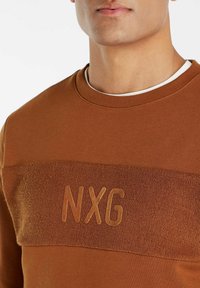 Bruine crewneck sweatshirt met textuurpaneel, featuring een reliëf "NXG" logo. Gemaakt van zachte stof, met gladde en getextureerde delen.