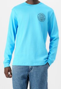 Camisa de manga larga azul claro, fabricada en algodón, con un cuello redondo y un logo circular negro en el lado izquierdo del pecho. Corte informal.