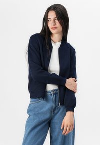 A young woman in a navy bomber jacket and white shirt stands with her arms crossed, gazing thoughtfully, set against a plain white background.
