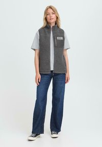 Femme aux cheveux blonds portant un gilet en polaire gris sur un t-shirt gris clair, un jean bleu à jambe large et des baskets noires, se tenant devant un fond uni.