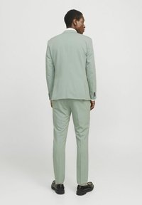 Traje verde claro con un corte entallado, solapa con muesca y una abertura trasera. Los pantalones tienen un corte recto. Zapatos negros completan el look.