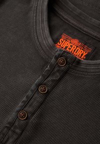 Camiseta negra de manga larga con textura acanalada, escote redondo y tres botones marrones. Presenta una etiqueta naranja "Superdry" en el interior del cuello.