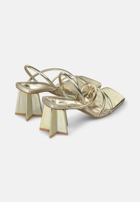 Goldene Riemchensandalen mit quadratischer Zehenform, ausgestattet mit metallischen Akzenten und einzigartigen skulpturalen Absätzen in Sternform mit einer cremefarbenen Oberfläche.
