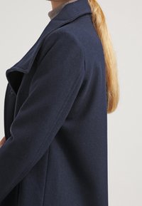 Cappotto in lana navy con una struttura sartoriale, dotato di un colletto a revers, tasche laterali e una texture liscia, abbinato a un leggero dolcevita.