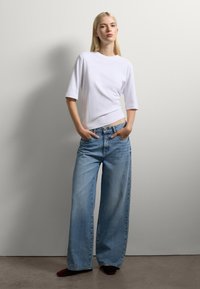 Donna bionda in piedi con le mani in tasca, che indossa un top bianco con maniche a gomito e jeans blu a gamba larga, contro uno sfondo di parete semplice.