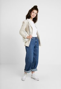 Beige blazer over en hvid t-shirt, højtaljede mørkeblå jeans med optrækkede manchetter og hvide sneakers; minimal hardware og rene linjer.