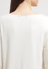 Maglietta a maniche lunghe di colore crema con vestibilità rilassata. Tessuto morbido, scollatura ampia e texture sottile. Nessun motivo visibile o dettagli in metallo.