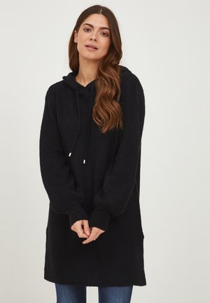 Svart ribbstickad hoodie-klänning med dragsko, långa ärmar, sidfickor och en avslappnad passform, parat med blå jeans.