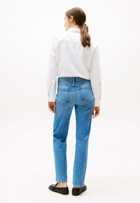 Jeans de mezclilla azul de pierna recta con una cintura de media altura, que cuentan con dos bolsillos traseros y un lavado sutil. Combinados con una camisa blanca de botones.