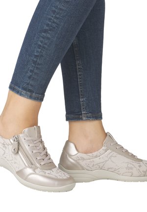 Baskets beiges avec une tige en tissu texturé et à motifs, agrémentées d'accents métalliques. Dotées d'une fermeture éclair latérale et d'un laçage, portées avec un jean bleu.